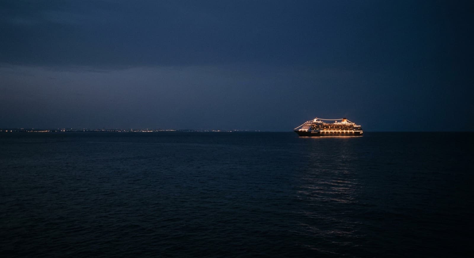 Un crucero iluminado en mar abierto, con la costa reducida a una línea de luces en el horizonte